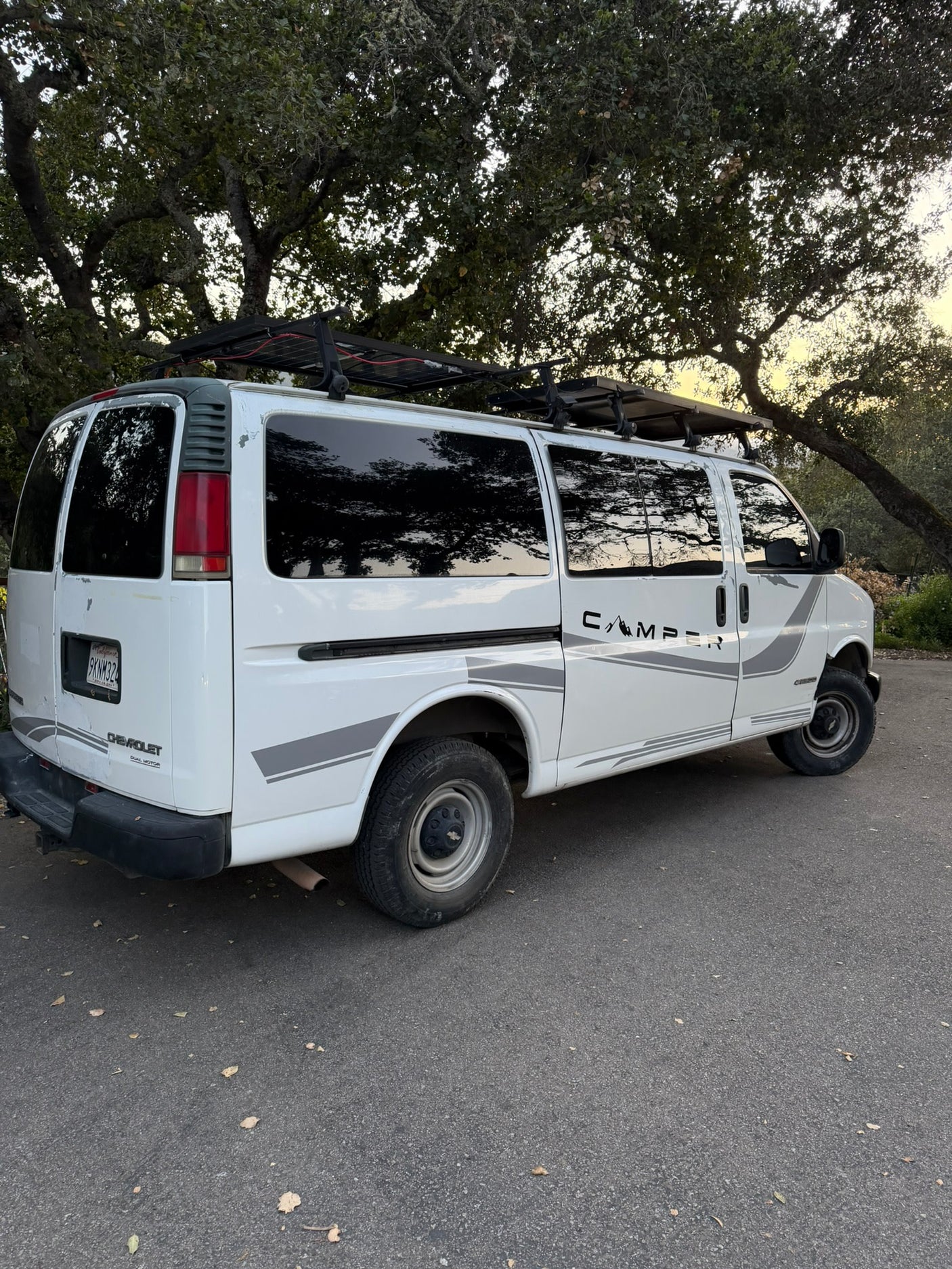 White Chevy Express camper van with rooftop solar panels and "CAMPER" decal, parked under trees. Ready for your rv rental adventure.
