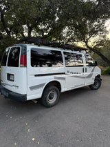 White Chevy Express camper van with rooftop solar panels and "CAMPER" decal, parked under trees. Ready for your rv rental adventure.