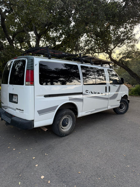 White Chevy Express camper van with rooftop solar panels and "CAMPER" decal, parked under trees. Ready for your rv rental adventure.