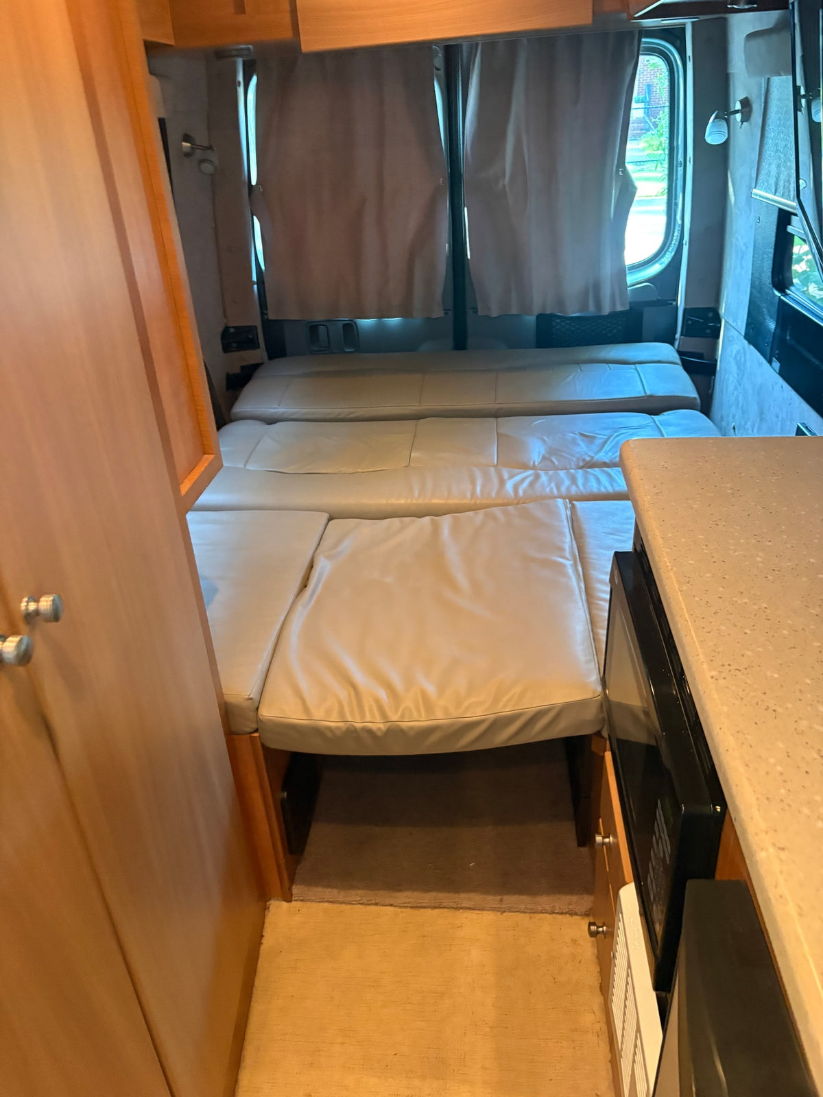 Compact RV rental interior with a light-colored, convertible bed/seating area, wooden cabinetry, and a kitchenette with a microwave. — photo 6