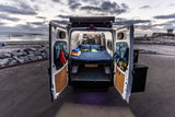 This RV rental camper van, parked beachside, reveals a cozy bed and organized interior through its open rear doors.