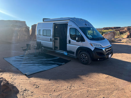 RV rental camper van parked in a sunny desert landscape with rock formations. Outdoor mat & chairs set up.