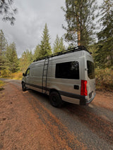 Overcast day with a silver camper van, an ideal **rv rental**, parked on a forest road covered in pine needles.