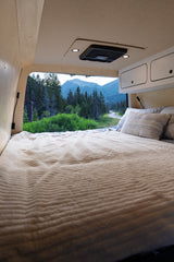 Inside an RV rental: a comfy bed with a soft blanket & pillows, overlooking a scenic mountain road, ideal for van life adventures.