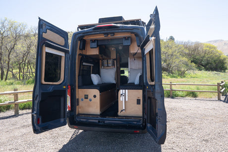 RV rental: Dark blue camper van with rear doors open, showcasing its customized wood interior, parked in a scenic outdoor area.