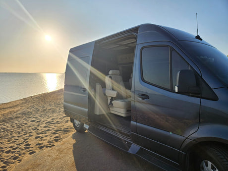 Luxurious RV rental parked on a sandy beach at sunset, side door open revealing comfortable interior seats.