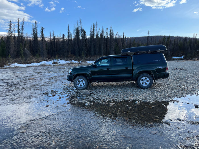 Dark green truck with rooftop tent on a rocky riverbed, perfect for an off-grid RV rental adventure. Mountains & forest view.