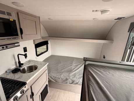 This rv rental features a compact interior with a kitchen (sink, stove, microwave), a bed with gingham bedding, and overhead storage.