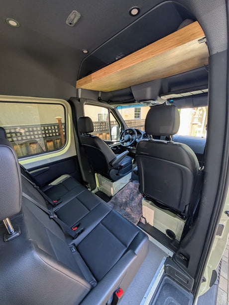 Interior of an RV rental van featuring multiple rows of black leather seating and a wooden overhead storage shelf.