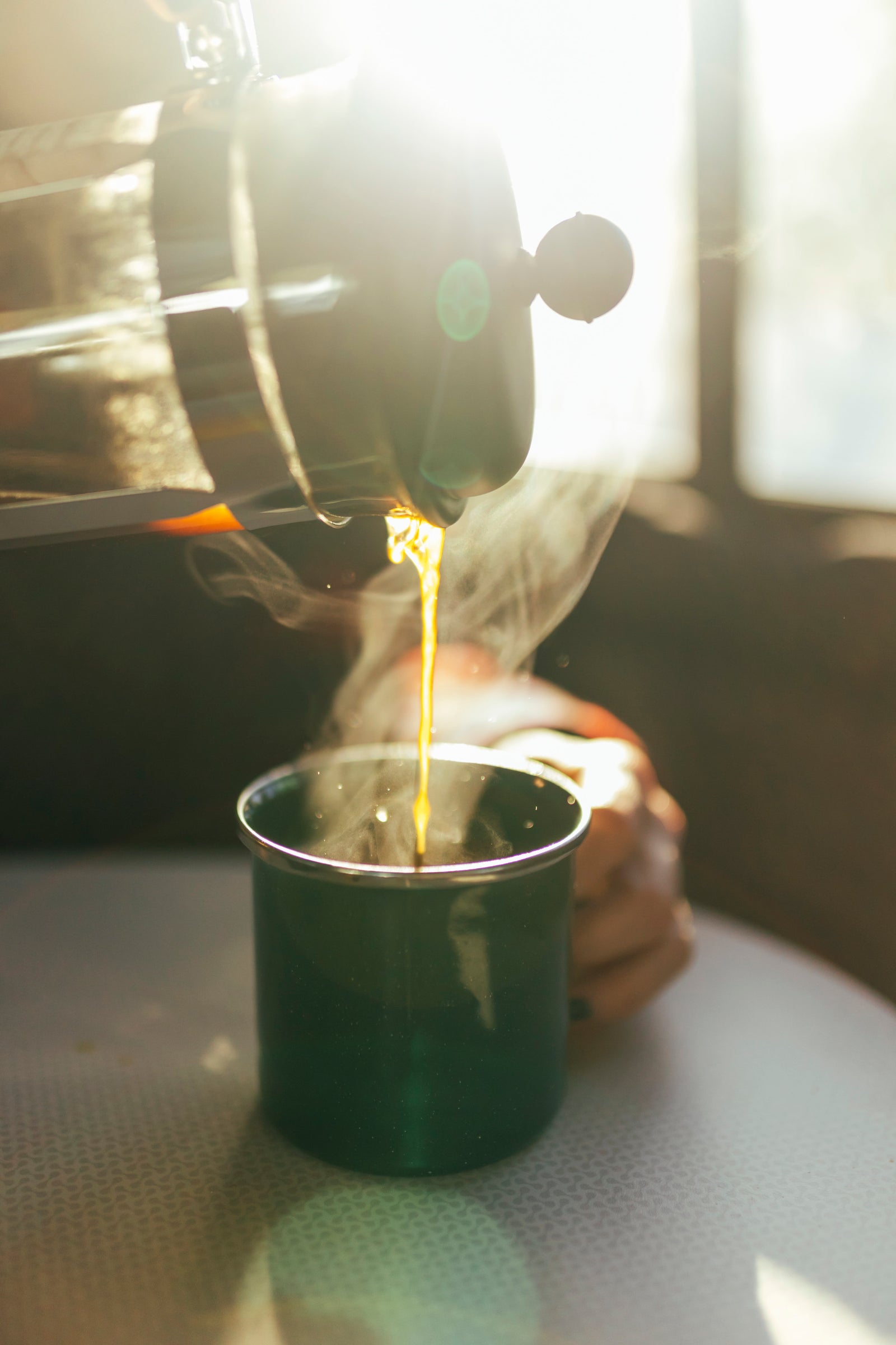 Person pouring steaming coffee from a French press into a green mug, bright morning inside an RV rental.