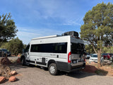 Silver Tellaro camper van rv rental parked on a gravel lot under a blue sky, surrounded by trees.