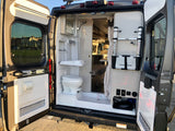 RV rental's rear doors open to reveal a compact bathroom with shower, toilet, and van interior.