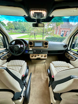 Luxury rv rental interior: Mercedes cockpit, plush cream quilted seats, and wooden dashboard trim with road view.