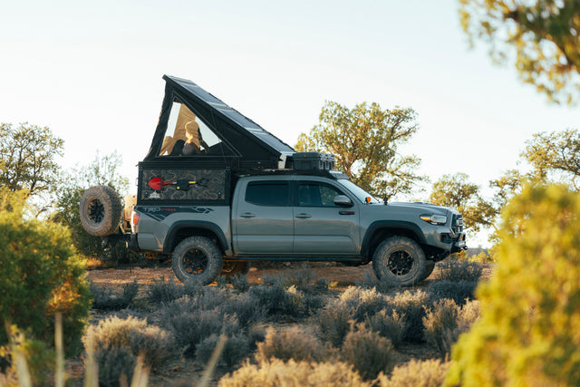 Explore off-road! This Toyota Tacoma overlanding truck camper RV rental is set in nature, with a person relaxing in its pop-top tent.