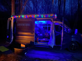 Adventure Rig RV rental van at night, illuminated by colorful string lights and a bright blue interior, parked for a cozy camp.