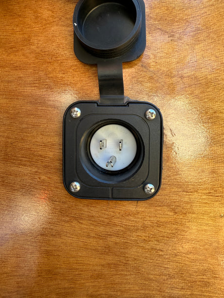 This RV rental's 15A/125V shore power inlet, with its protective cover open, is ready for hookup on a wooden exterior.