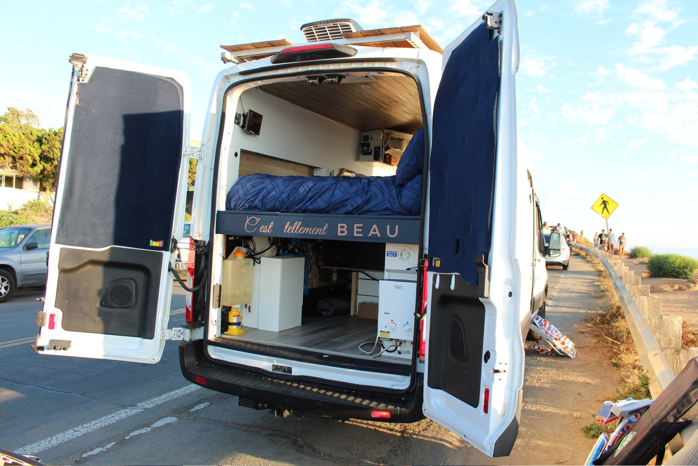 Open rear doors of a customized white camper van, showcasing its cozy bed, wood interior, and utilities, perfect for an rv rental.