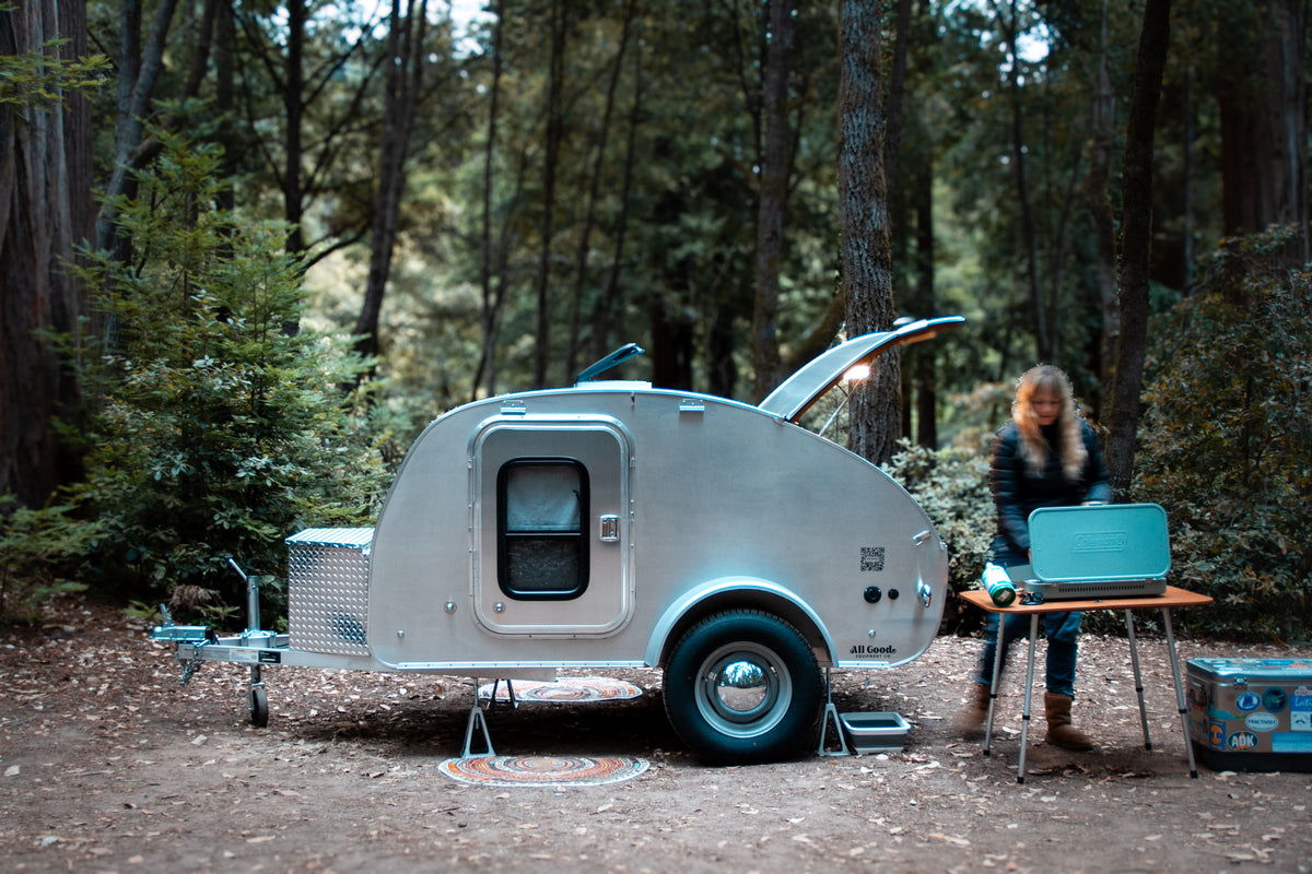 Teardrop RV rental at a forest campsite, with a person setting up a Coleman stove for cooking. Ideal for outdoor adventures.