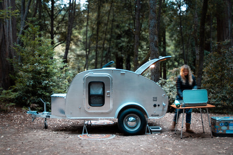 Teardrop RV rental at a forest campsite, with a person setting up a Coleman stove for cooking. Ideal for outdoor adventures.