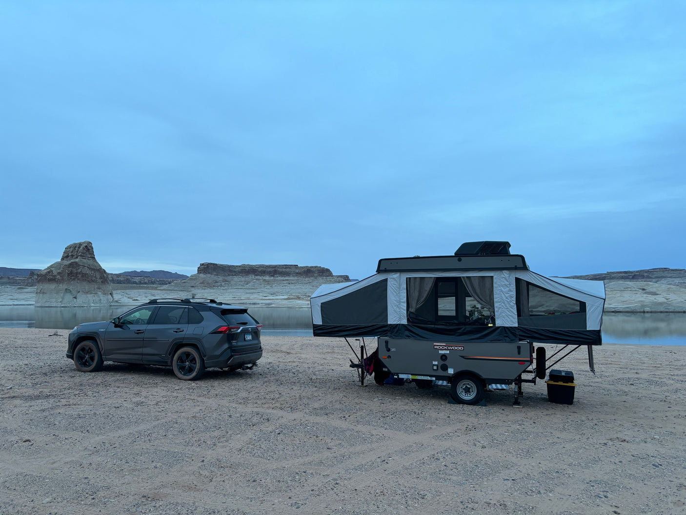 An RV rental pop-up camper set up on a sandy lake beach next to an SUV, with large rock formations in the background.