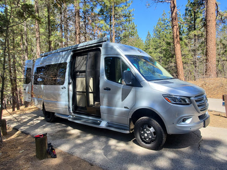 Silver Mercedes Sprinter RV rental parked in a sunny forest campsite, side door open. Shoes on a stump nearby.