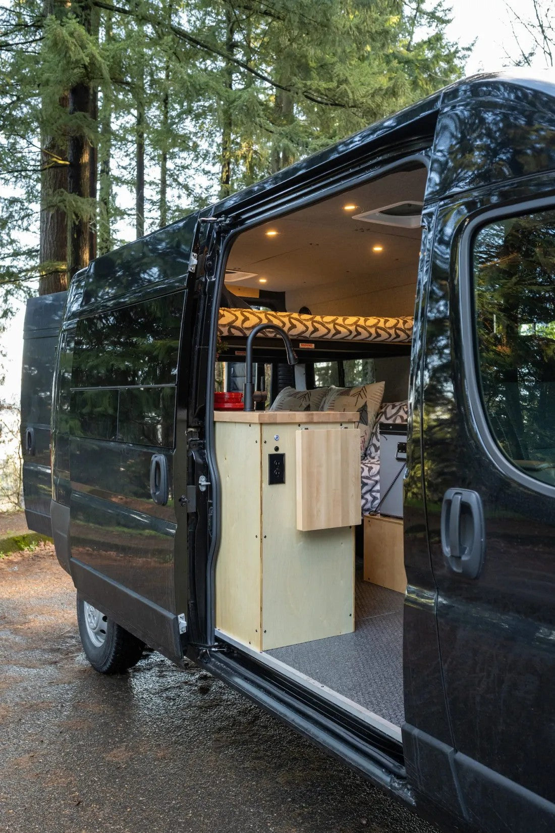 Open black camper van for RV rental, revealing a cozy interior with kitchen, bed, and modern amenities amidst tall trees.