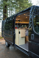 Open black camper van for RV rental, revealing a cozy interior with kitchen, bed, and modern amenities amidst tall trees.