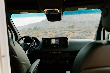 Inside a Mercedes-Benz rv rental van, the dash and screen show a scenic desert road trip view with mountains in the distance.