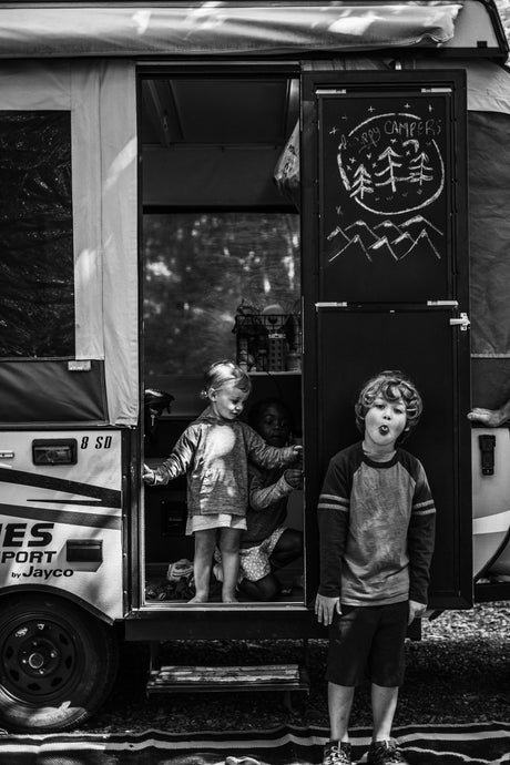 An RV rental brings joy: Kids play around a pop-up camper, with one boy making a silly face outside. "Happy Campers" chalkboard visible.