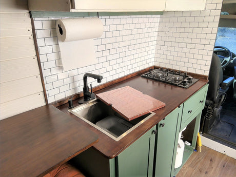 This RV rental kitchen boasts dark counters, white subway tile, sink, stovetop, and green cabinets for comfy travels.