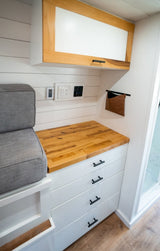 Modern RV rental interior: white shiplap walls, wood countertop, storage drawers, overhead cabinet, and comfy seating.