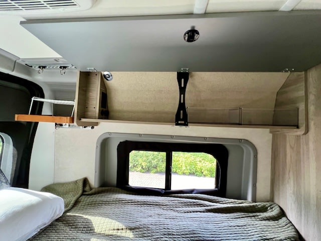 RV rental interior showcasing a bed, open overhead cabinet with storage solutions, and a window view.