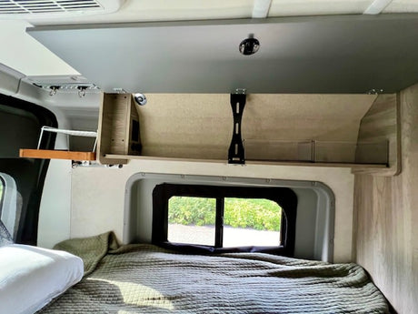 RV rental interior showcasing a bed, open overhead cabinet with storage solutions, and a window view.