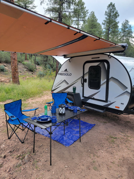 This RV rental teardrop camper is set up for camping with an extended awning, chairs, and table at a peaceful campsite.