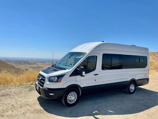 White Ford Transit RV rental van parked on a dirt road, overlooking dry hills and a distant town under a clear blue sky.
