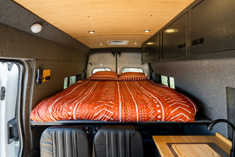 Cozy RV rental interior, showcasing a comfy bed with orange bedding, wooden ceiling, and a compact kitchen setup.