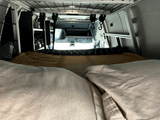 Comfortable bed inside an RV rental, facing the vehicle's cabin with overhead storage and lighting.