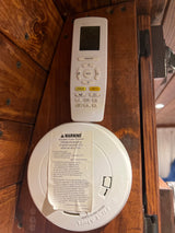An RV rental interior shows a First Alert CO/smoke alarm with warning label below an air conditioner remote on a wooden wall.