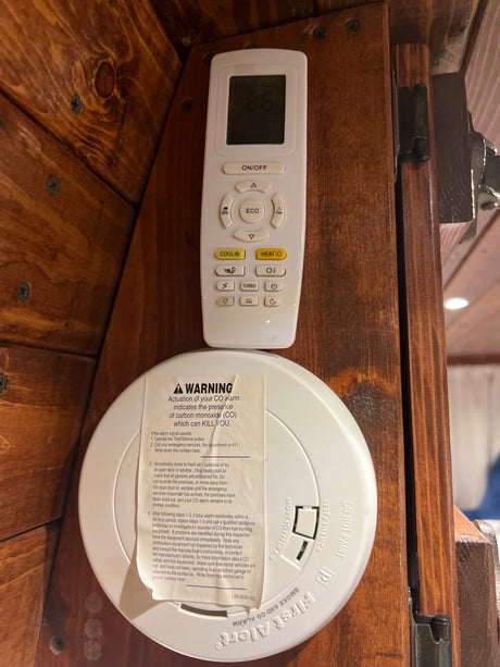 An RV rental interior shows a First Alert CO/smoke alarm with warning label below an air conditioner remote on a wooden wall.