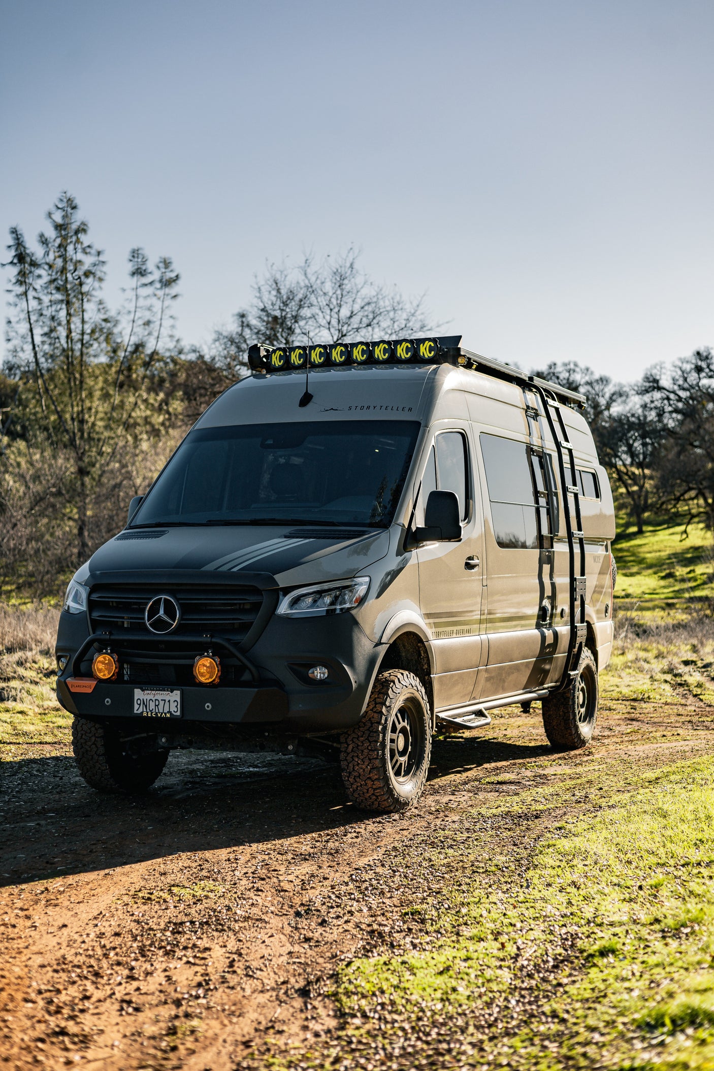 An overland Mercedes Sprinter camper, ideal for an rv rental, with off-road tires, KC lights, and a side ladder on a dirt trail.