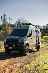 An overland Mercedes Sprinter camper, ideal for an rv rental, with off-road tires, KC lights, and a side ladder on a dirt trail.