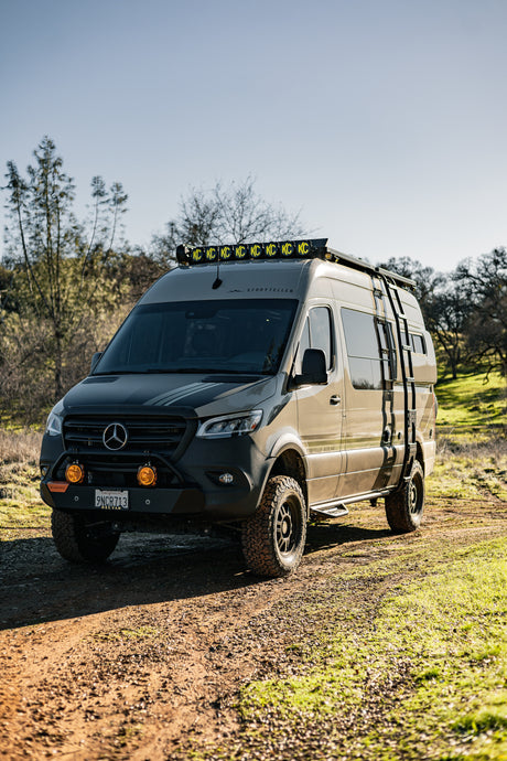 An overland Mercedes Sprinter camper, ideal for an rv rental, with off-road tires, KC lights, and a side ladder on a dirt trail.