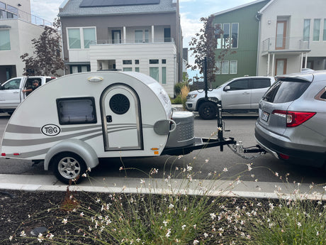 White T@G teardrop RV rental hitched to an SUV, parked street-side in a modern residential area.