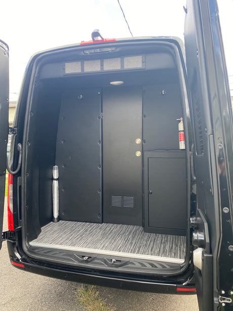 Spacious custom black van interior for RV rental. Features specialized paneling, flooring, storage, & safety equipment.