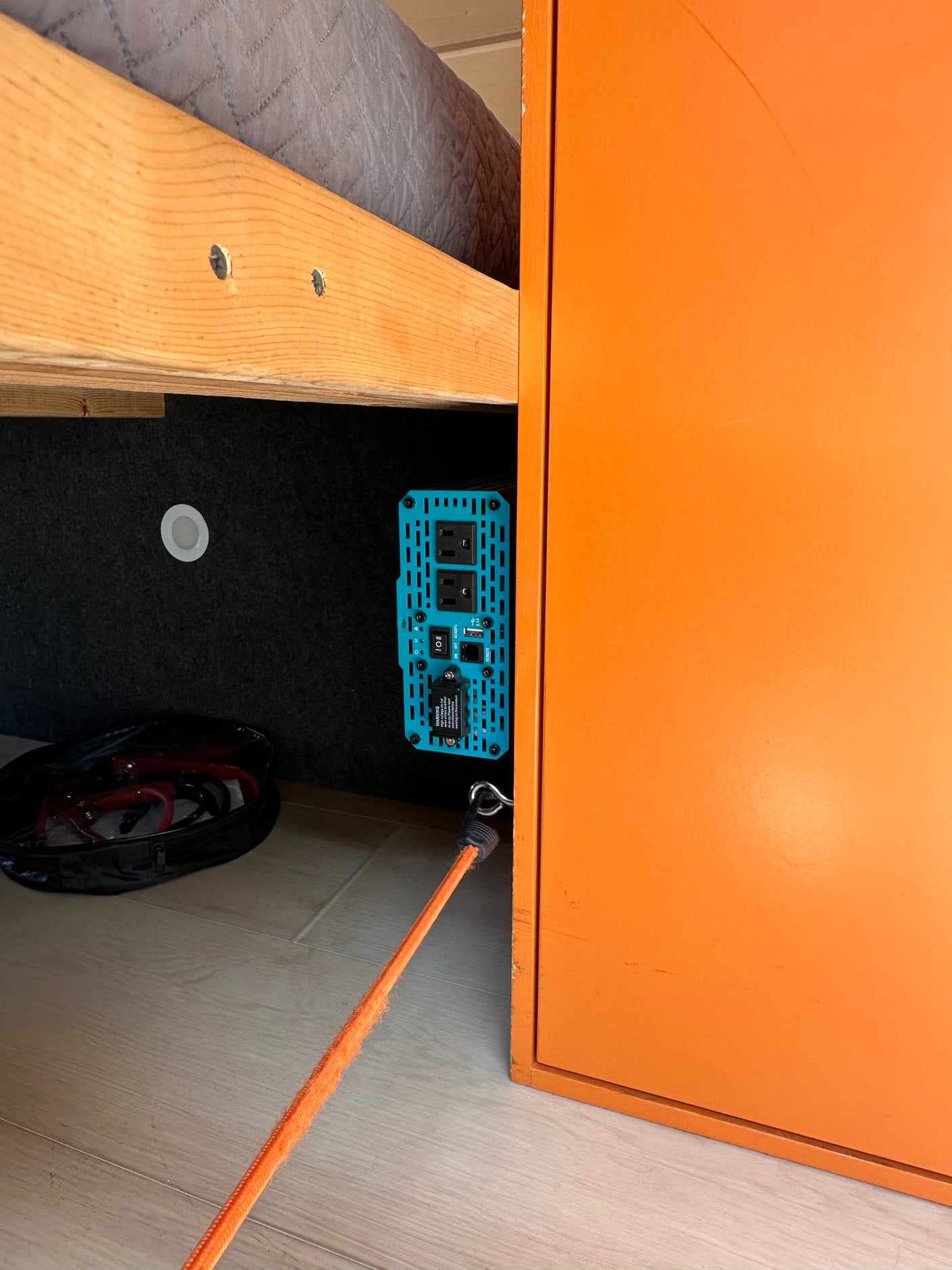 Inside an RV rental: a blue power inverter, light, and cable bag under a wooden bed frame, beside an orange cabinet.
