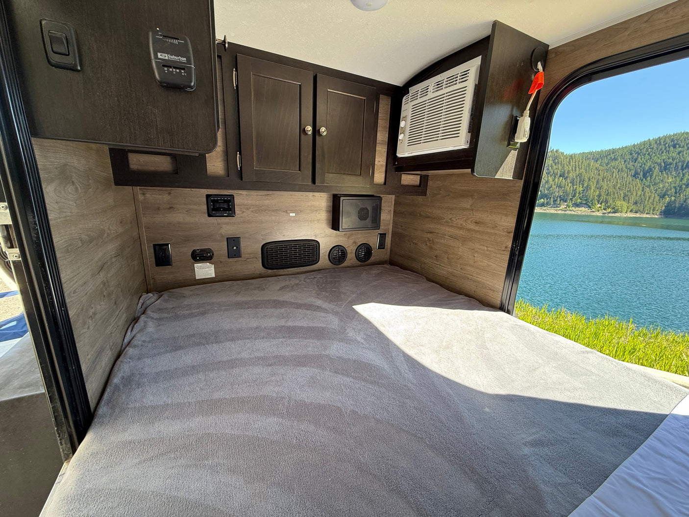 Inside a comfy RV rental: cozy bed, control panels, AC unit, & scenic lake view through the window overlooking mountains.