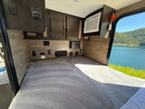 Inside a comfy RV rental: cozy bed, control panels, AC unit, & scenic lake view through the window overlooking mountains.
