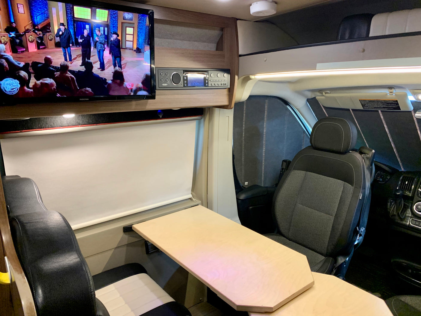 Inside an RV rental, a TV plays in the lounge area with a dining table and front driver's seat visible.
