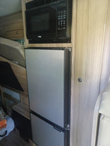 RV rental kitchen featuring a black microwave and two light-grey privacy shades integrated into light wood cabinetry, showcasing a compact interior.