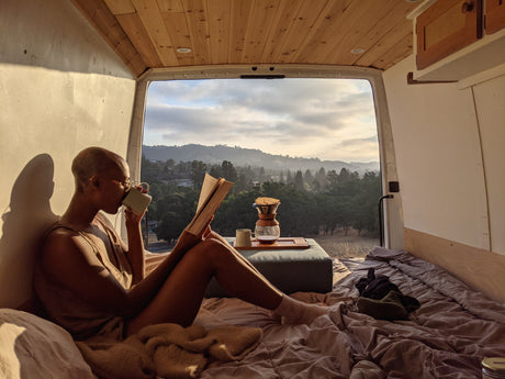 A person enjoys coffee & a book in an rv rental, with open back doors showing misty mountains & trees.